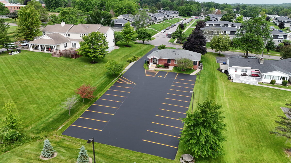 This overhead view captures a freshly sealed commercial parking lot surrounding a multi-story building, treated with a premium, sand-fortified rubberized asphalt emulsion. Engineered for long-lasting performance, this polymer-enhanced sealer reconditions and texturizes aged pavement while forming a waterproof barrier resistant to oil and gas damage. The result is a rich satin black surface with bright, well-defined striping. Ideal for high-traffic environments, it offers a two-coat coverage rate of 30 to 40 sq. ft. per gallon and enhances both durability and aesthetics.